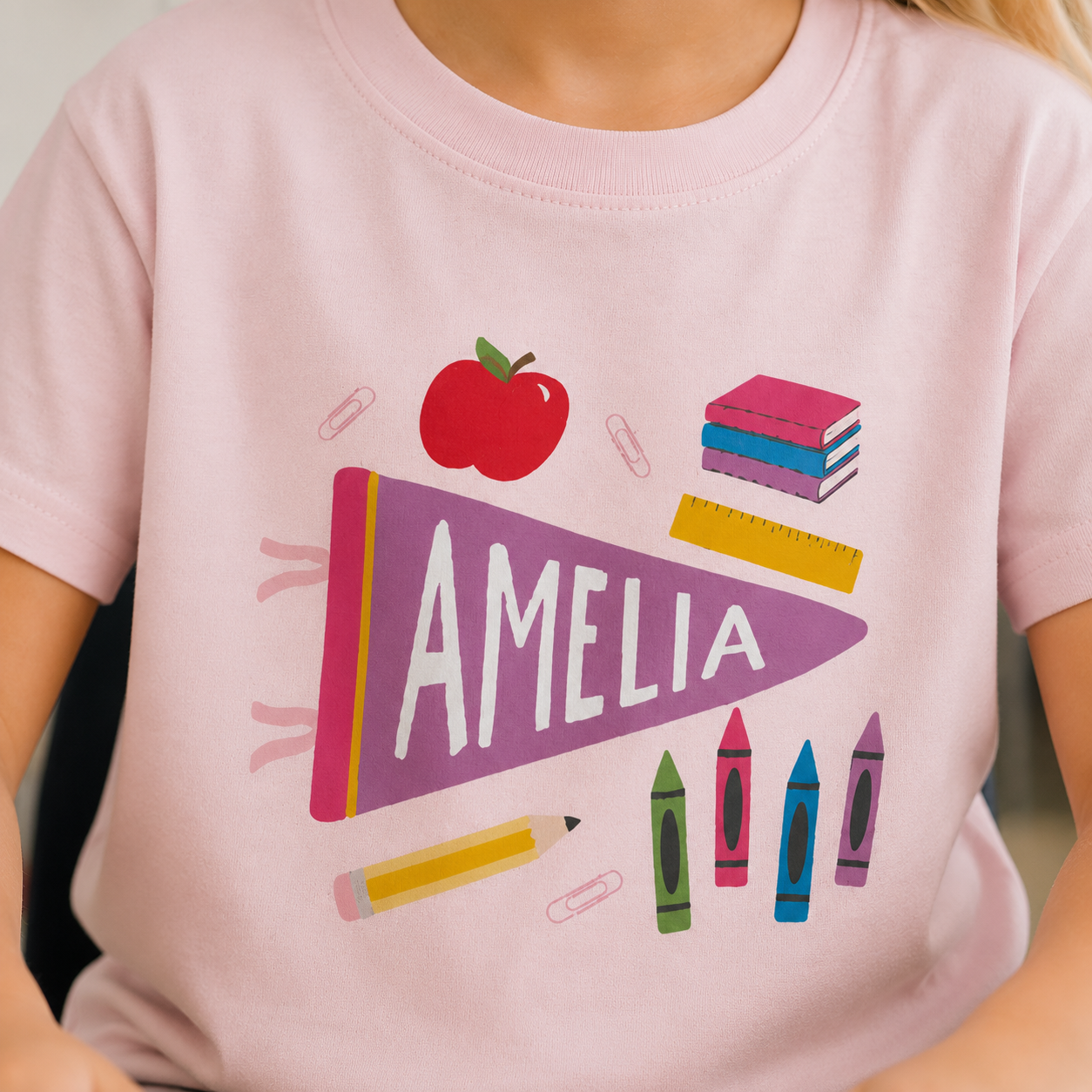 A close-up of a child wearing a light pink t-shirt with a colorful school-themed graphic. The graphic features a purple pennant with the name 'AMELIA' in white, surrounded by school supplies including a red apple, pink and blue books, a yellow pencil, and crayons.