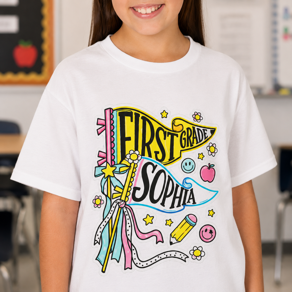 A child wearing a white short-sleeve graphic T-shirt with colorful text reading 'FIRST GRADE' and 'SOPHIA' over a whimsical school-themed illustration, photographed indoors with a blurred classroom background.