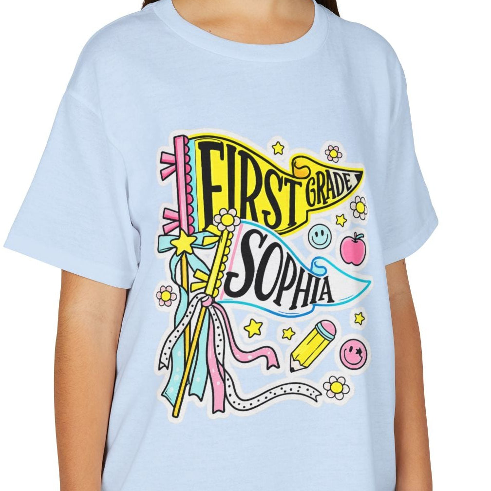 A young girl smiling while wearing a light blue crewneck t-shirt with a colorful 'First Banana Sophia' graphic on the front, paired with khaki cargo pants, against a plain white background.