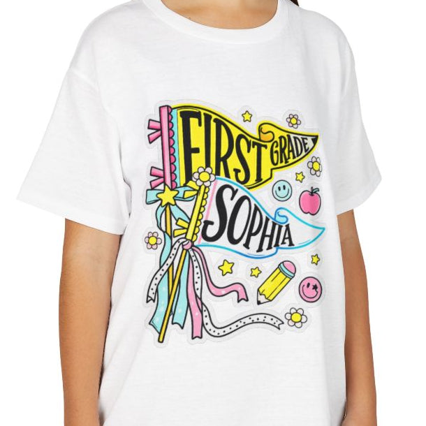 A young girl smiling while wearing a white short-sleeve graphic t-shirt with a colorful 'First Star Sophia' design, paired with khaki cargo pants, against a solid white background.