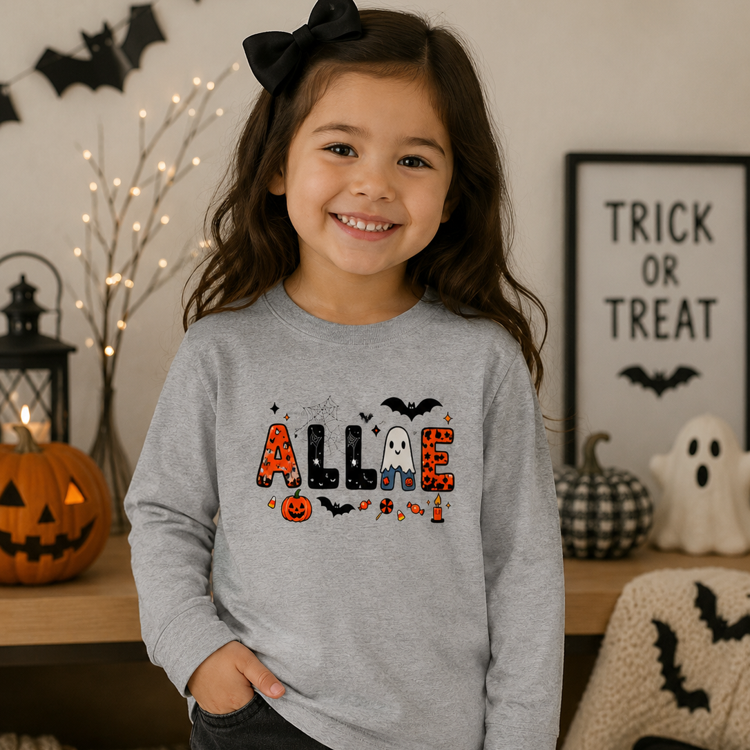A young girl with a black bow in her hair smiles while wearing a light gray long-sleeve sweatshirt with a 'ALLIE' Halloween graphic. The background shows a cozy, decorated Halloween scene with pumpkins, a ghost, and a 'Trick or Treat' sign.