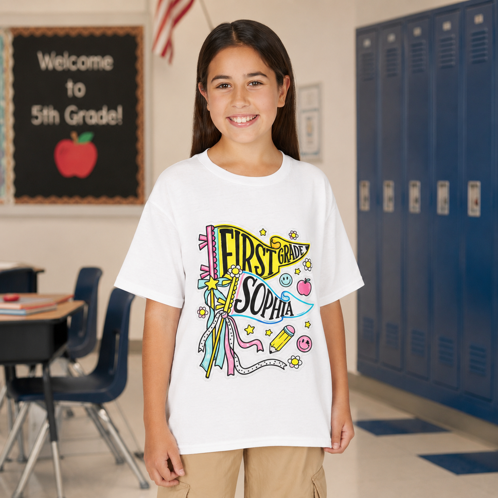 A young girl stands in a school hallway wearing a white graphic t-shirt with a colorful 'First Grade Sophia' banner design, paired with khaki cargo pants.