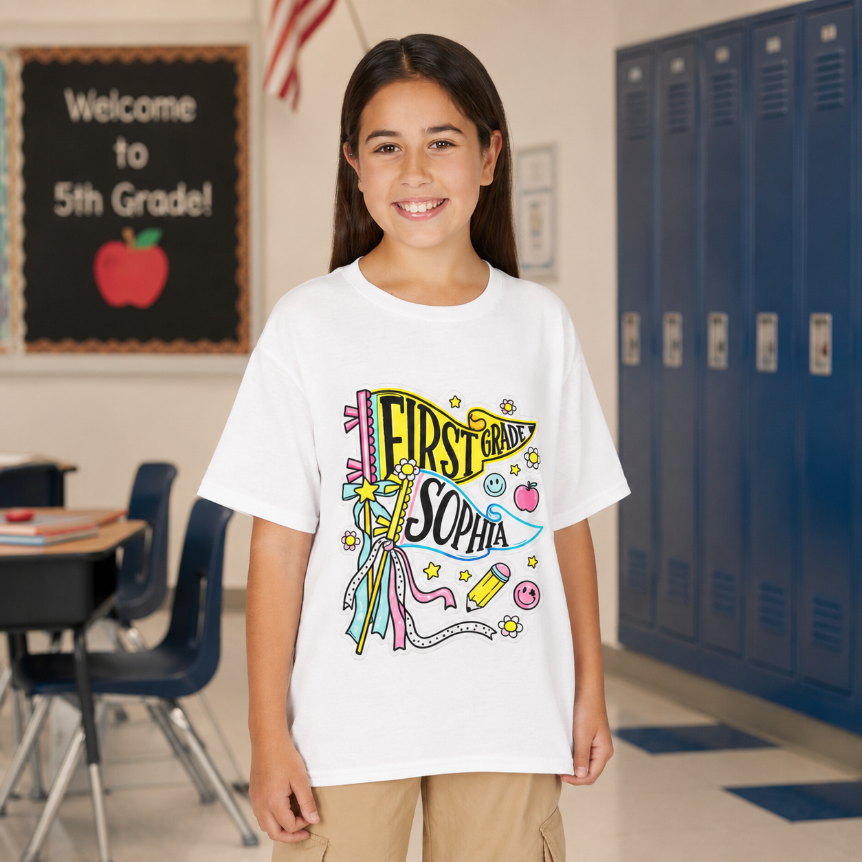 A young girl stands in a school hallway wearing a white graphic t-shirt with a colorful 'First Grade Sophia' banner design, paired with khaki cargo pants.