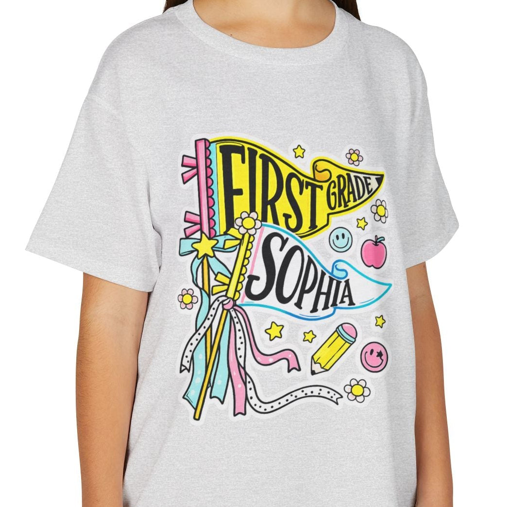 A young girl smiling while wearing a light gray heather t-shirt with a colorful 'First Banana Sophia' graphic on the front, paired with khaki cargo pants, against a plain white background.
