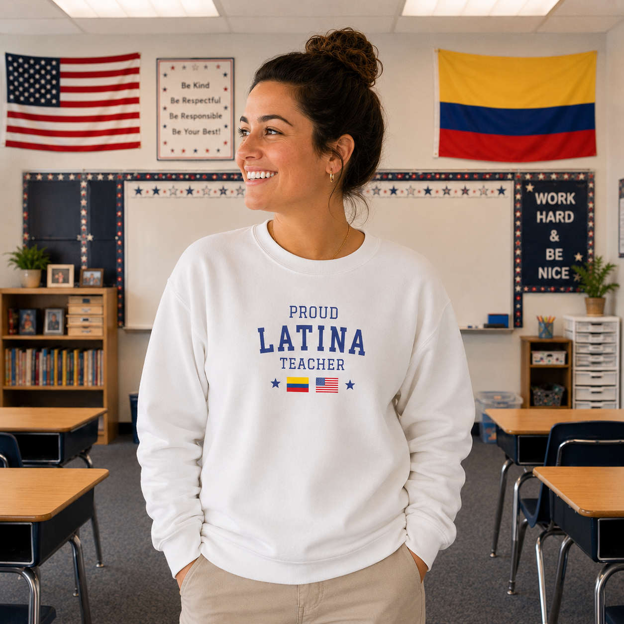 A woman stands in a classroom wearing a white crewneck sweatshirt with 'PROUD LATINA TEACHER' printed in navy blue, along with small flag graphics. The background shows desks, a whiteboard, and educational posters.