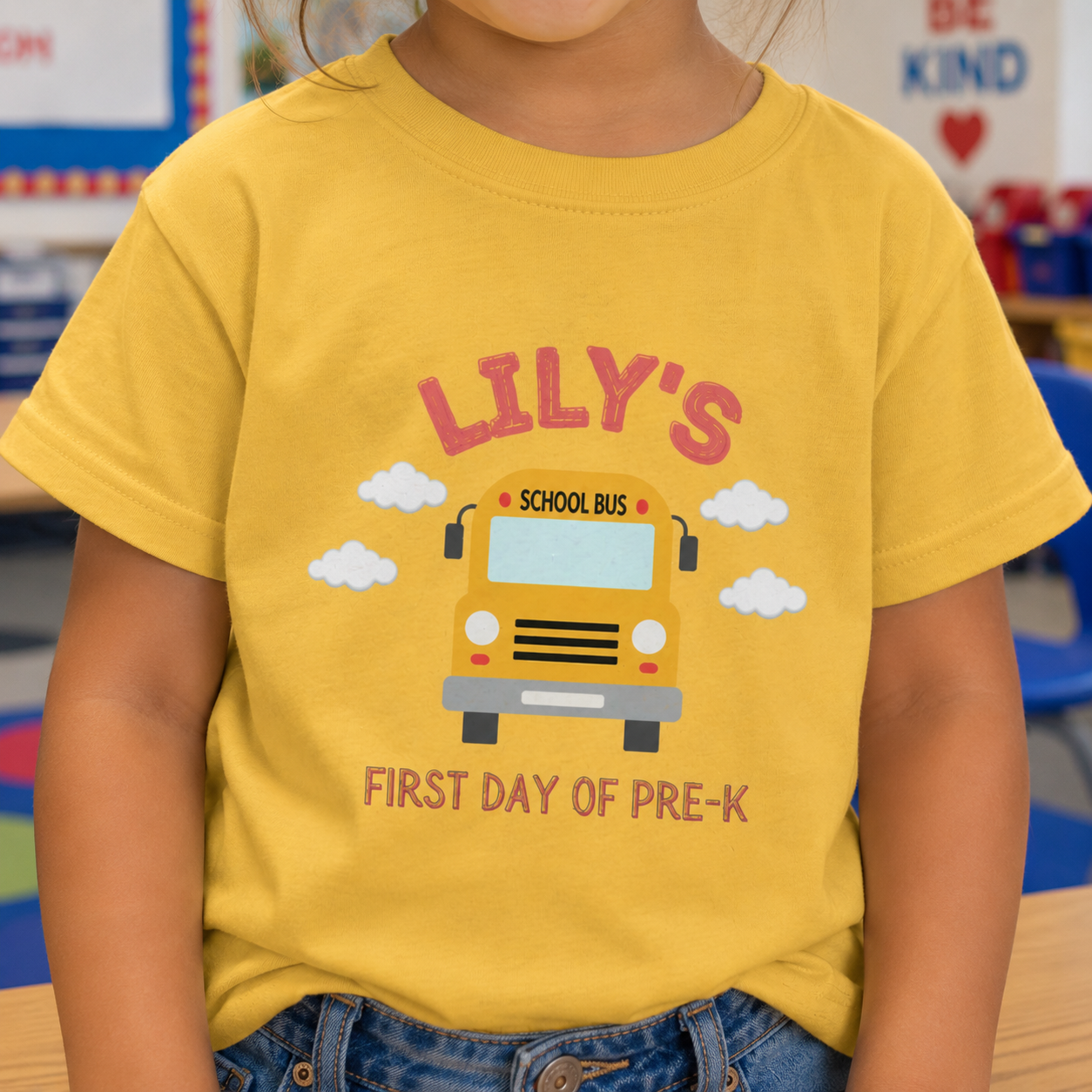 Girl wearing a yellow personalized "Lily's School Bus First Day of Pre-K" tee in a classroom