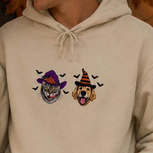 A man and a woman wearing matching cream-colored hoodies with a Halloween-themed graphic of a witch cat and a dog wearing a hat on the chest, posed in a cozy, decorated Halloween setting with pumpkins and string lights.