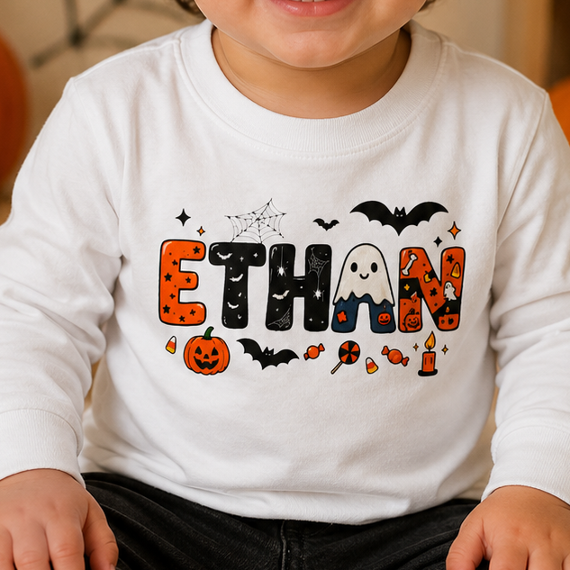 A smiling toddler sits cross-legged on a beige carpet in a Halloween-themed room, wearing a white long-sleeve shirt with a colorful 'ETHAN' graphic featuring bats, pumpkins, ghosts, and candy, paired with dark jeans and white sneakers.