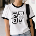 A woman wearing a white ringer t-shirt with black trim and a black graphic print that reads 'in my six seven era'. She is standing in a school hallway with lockers on the right.