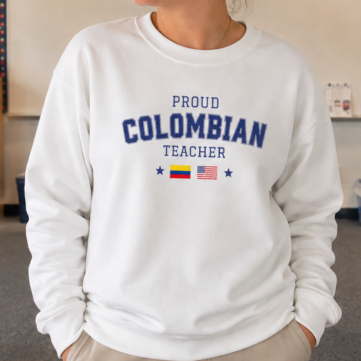 A woman stands in a classroom wearing a white sweatshirt with 'PROUD COLOMBIAN TEACHER' printed in navy blue text, along with small Colombian and American flag graphics. The background shows a classroom with desks, flags, and educational posters.