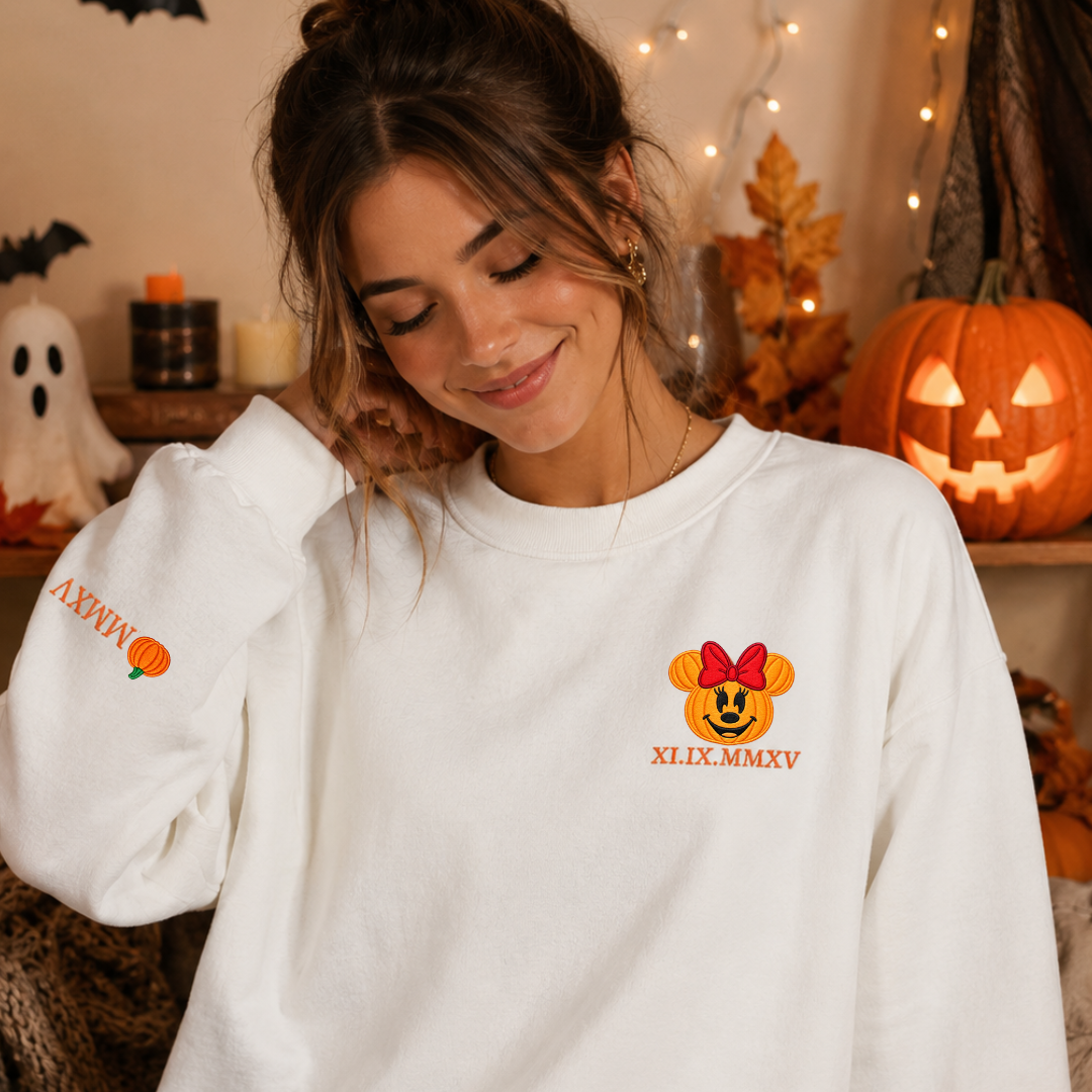 A woman wearing a white crewneck sweatshirt with a custom orange pumpkin graphic and 'ANVIL' text on the sleeve, posed in a cozy Halloween-themed room with pumpkins and fairy lights.