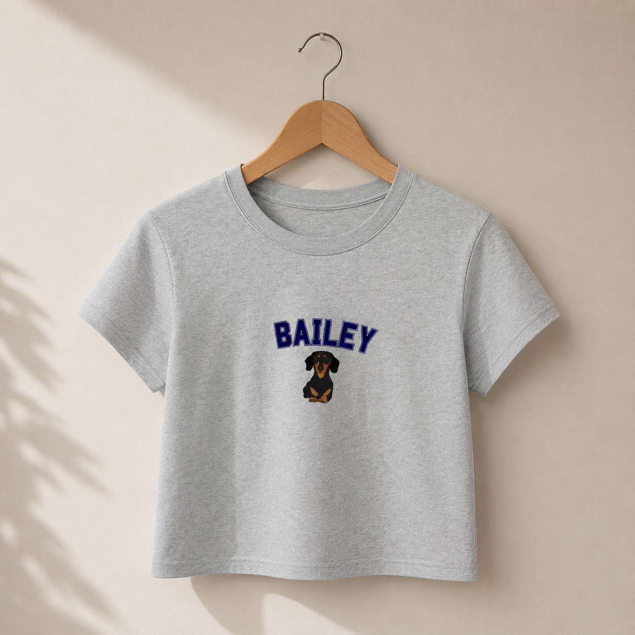 Gray t-shirt with 'BAILEY' and a dachshund graphic on a wooden hanger against a beige background