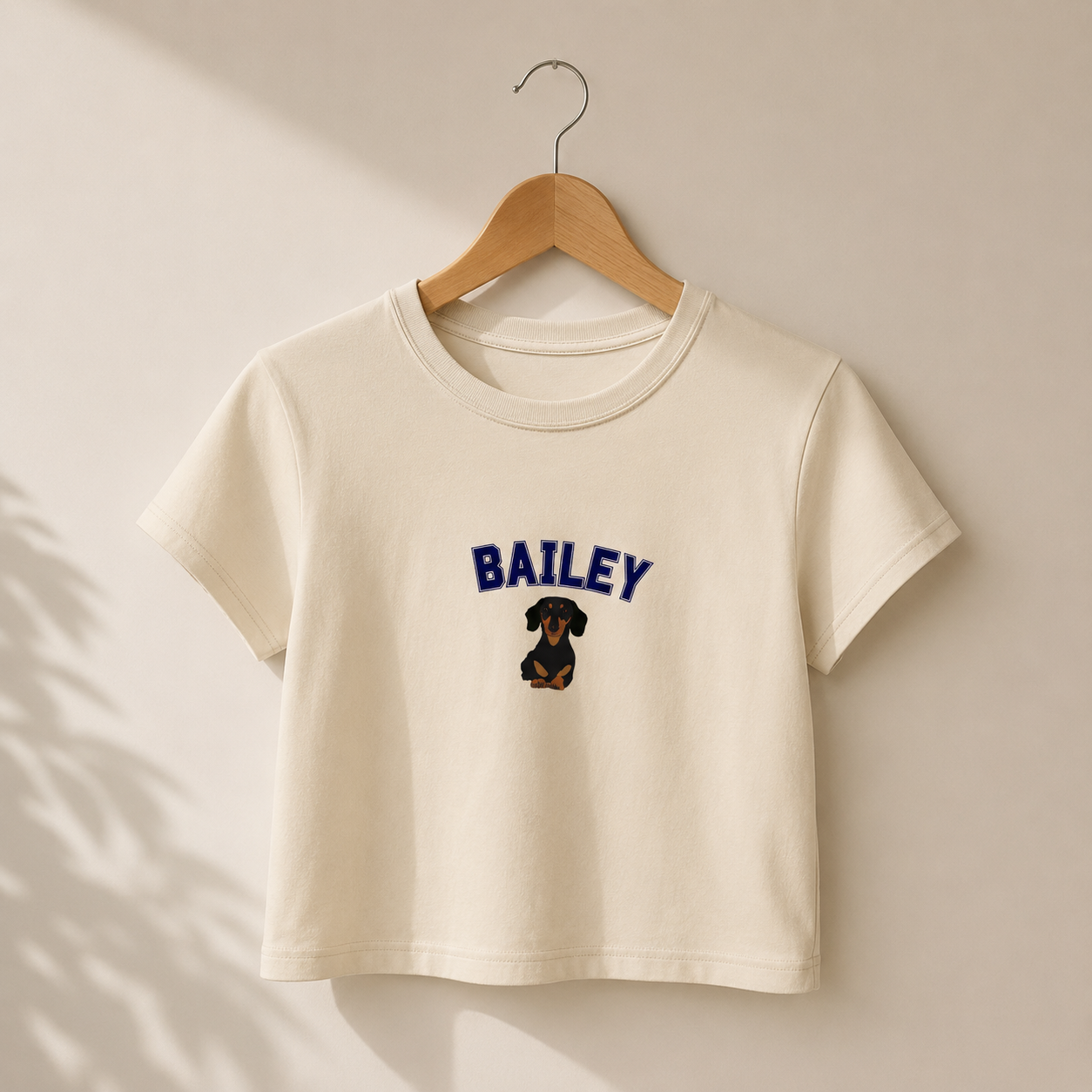 Beige t-shirt with 'BAILEY' and a dog graphic on a wooden hanger against a beige wall.