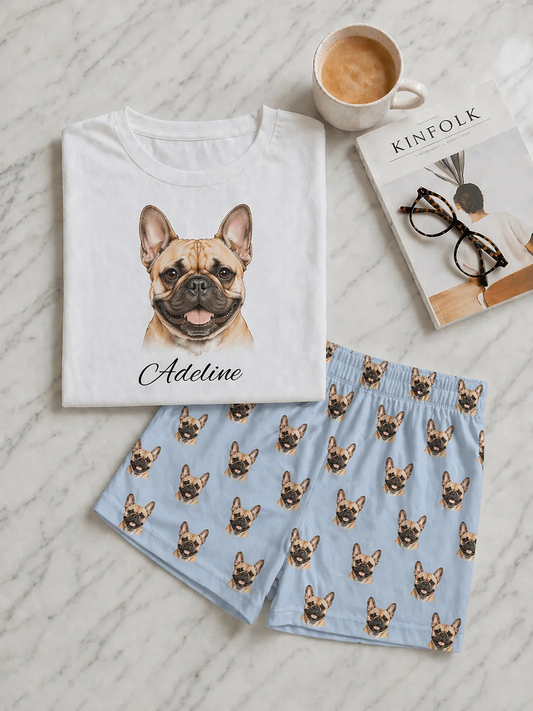 White t-shirt with a dog illustration and text, blue shorts with dog pattern, cup of coffee, and book on a textured surface.