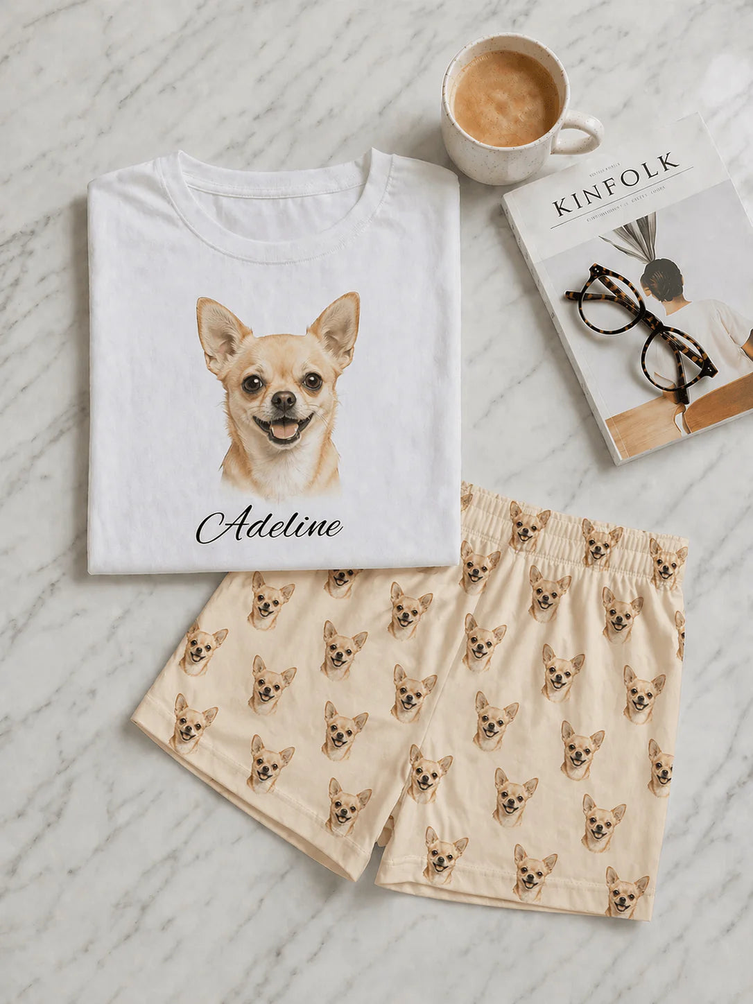 White t-shirt with a dog illustration and name, beige shorts with dog pattern, cup of coffee, and book on a light wooden surface.