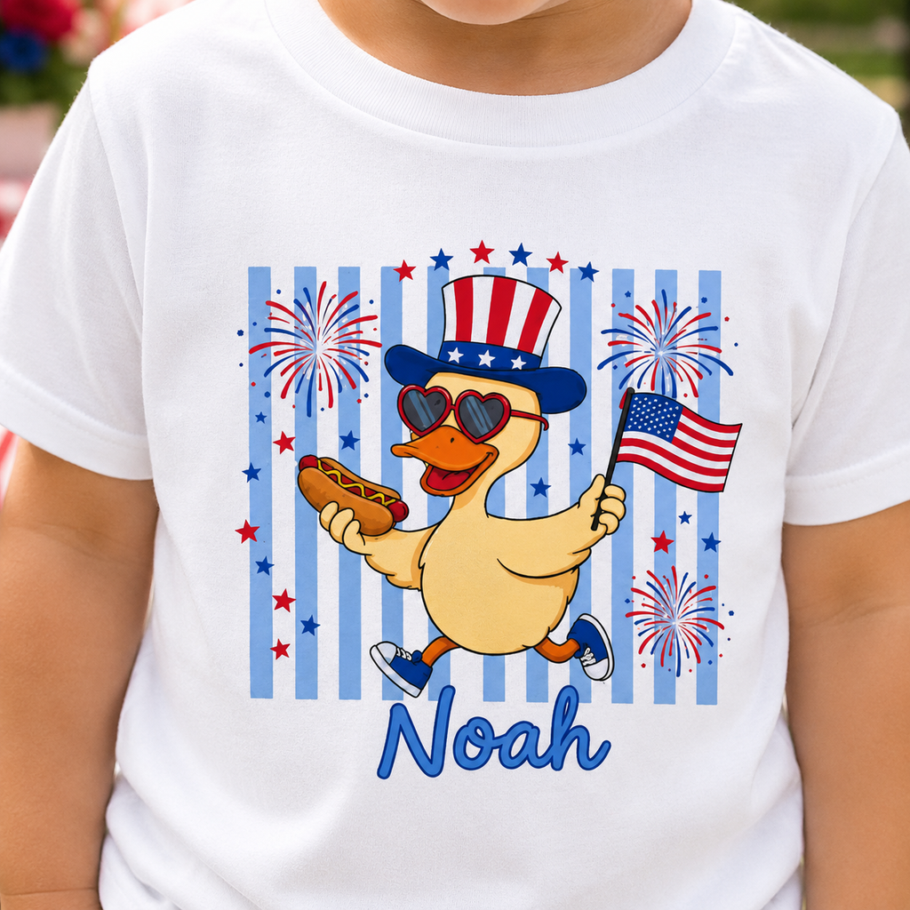 White t-shirt with a cartoon duck holding an American flag and a hot dog, surrounded by fireworks, with 'Noah' printed below.