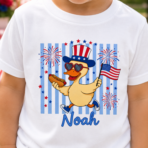 White t-shirt with a cartoon duck holding an American flag and a hot dog, surrounded by fireworks, with 'Noah' printed below.