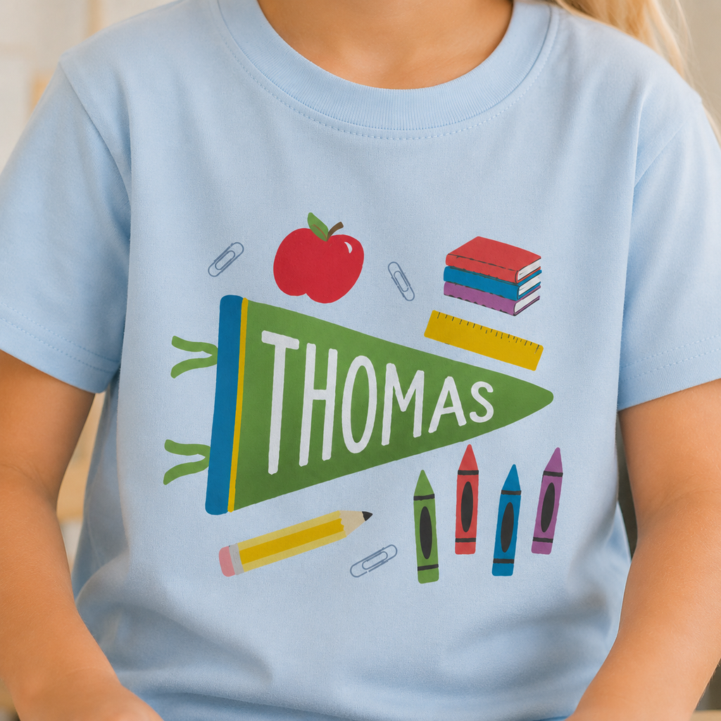 A child wearing a light blue t-shirt with a colorful school-themed graphic featuring a green pennant that says 'THOMAS' in white, surrounded by an apple, books, crayons, and paperclips.