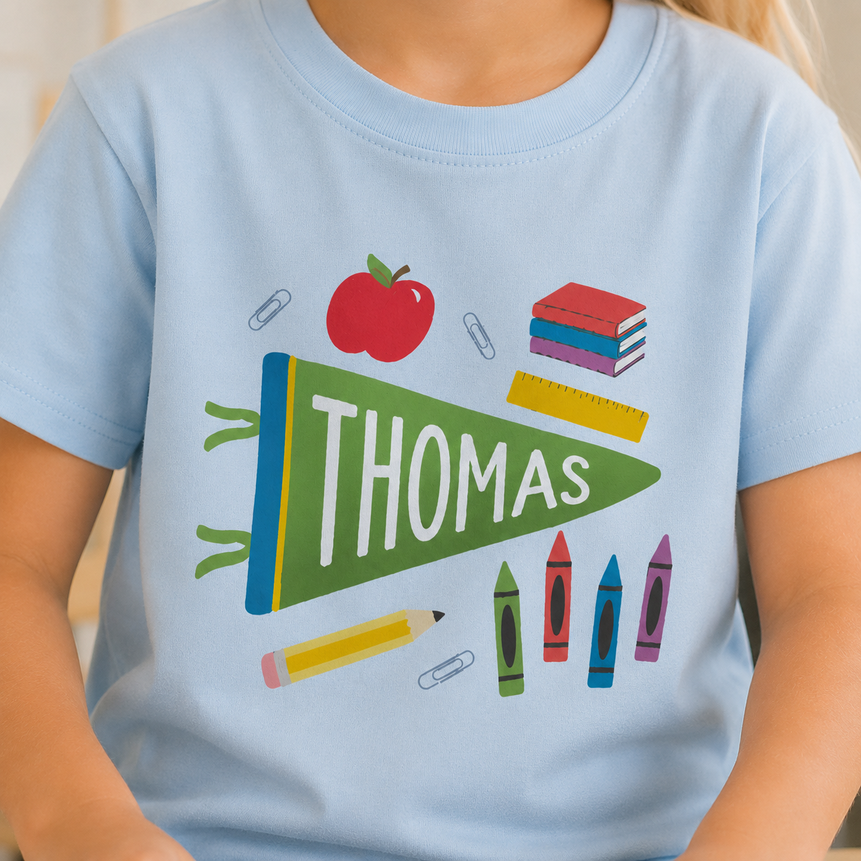 A child wearing a light blue t-shirt with a colorful school-themed graphic featuring a green pennant that says 'THOMAS' in white, surrounded by an apple, books, crayons, and paperclips.