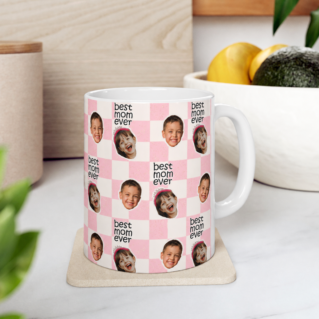 Mug with 'best mom ever' text and children's faces on a pink checkered background, placed on a table.