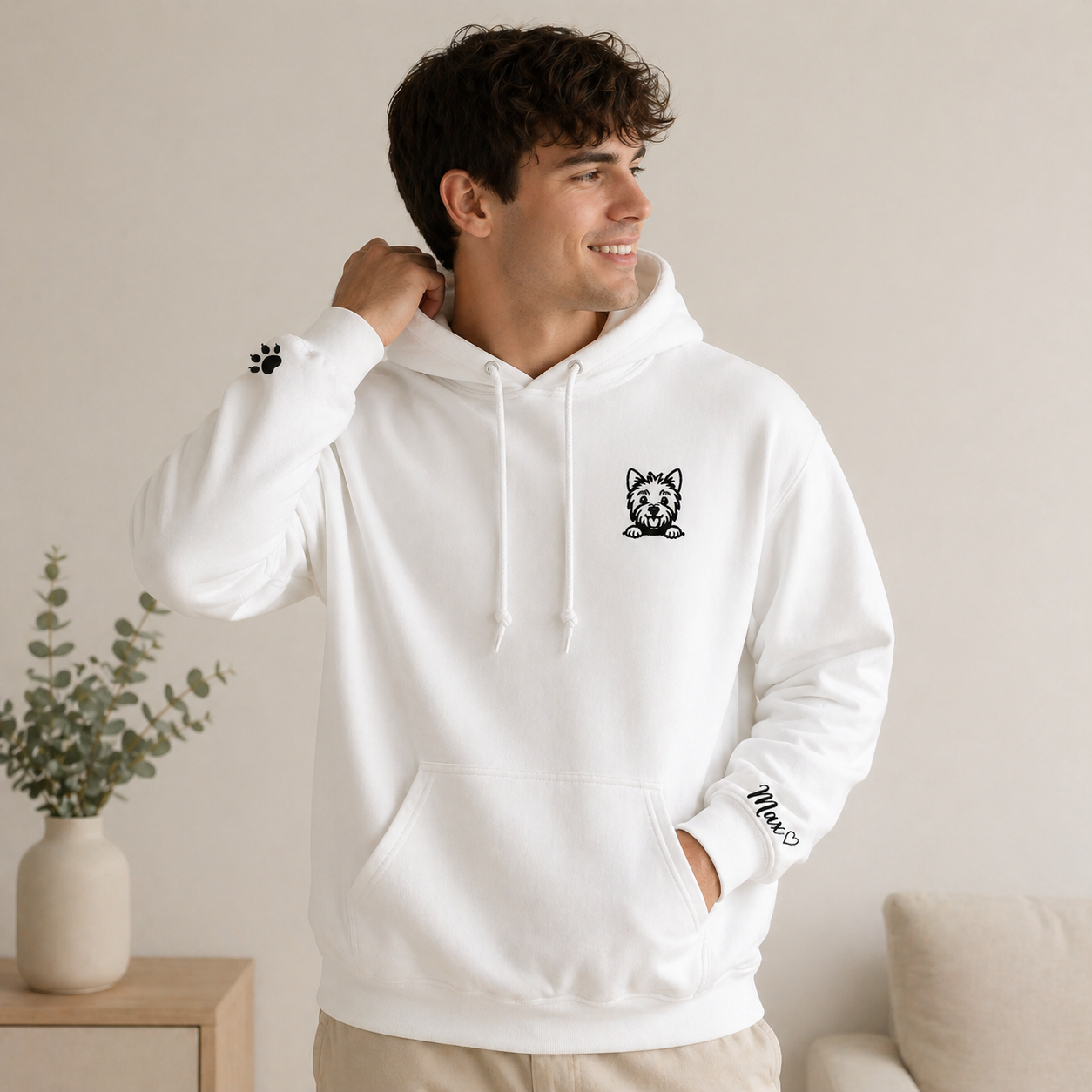 Man wearing a white hoodie with a logo, standing in a room with a plant and a couch.