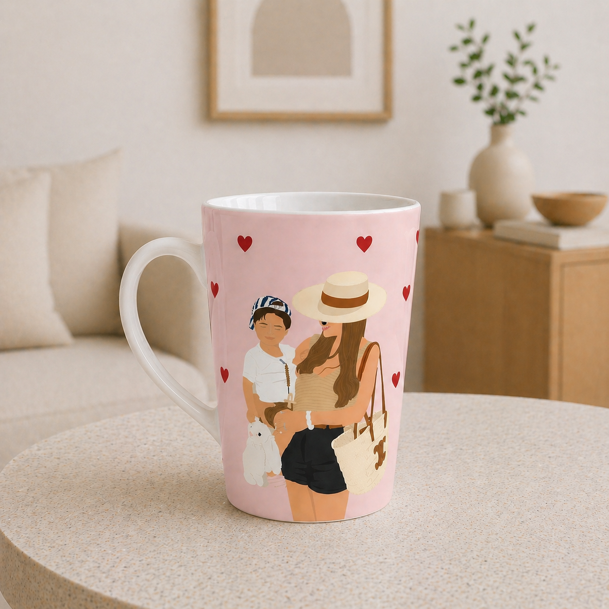 Pink mug with a design of a woman and child holding a dog, surrounded by hearts, on a table in a cozy living room.