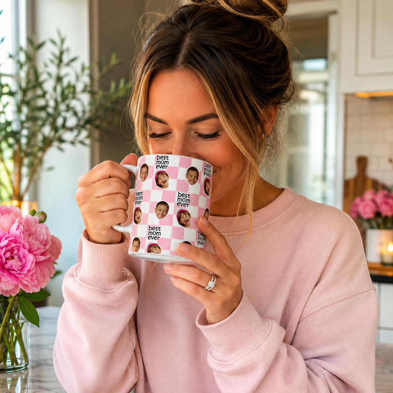 Woman in a pink sweater holding a mug with a 'best mom ever' design in a kitchen.