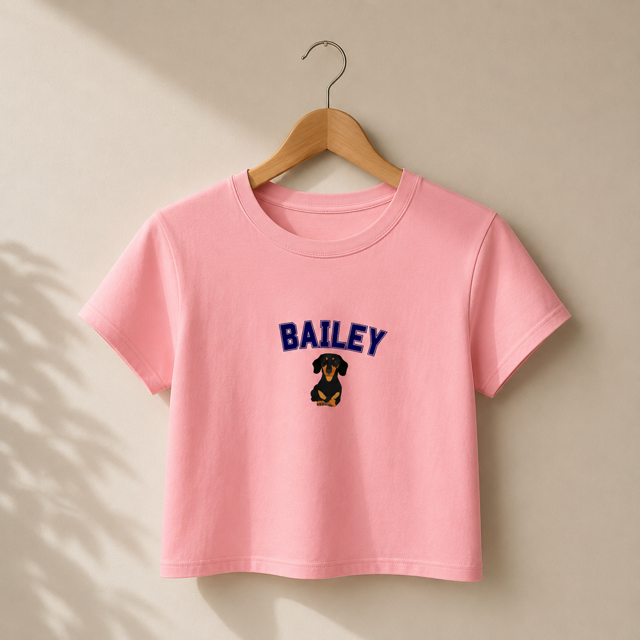 Pink t-shirt with 'BAILEY' and a dog graphic on a wooden hanger against a beige background